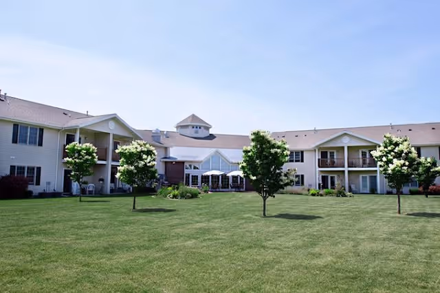 A large, two-story senior living facility building with beige siding and a brown roof, surrounded by a well-maintained green lawn with several small trees and shrubs under a clear blue sky.
