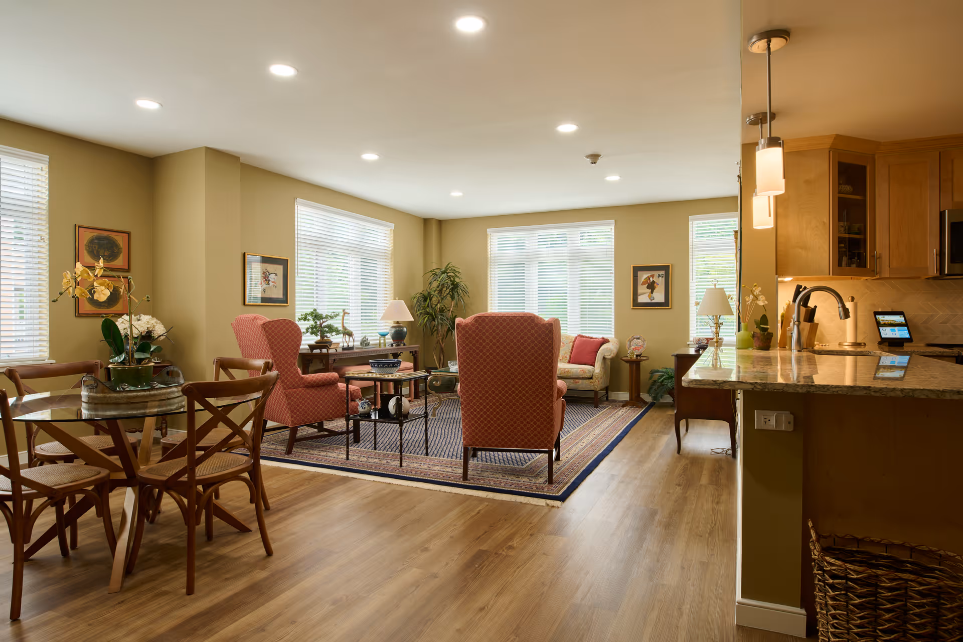 Well-lit communal living area with armchairs around a rug, a dining table, and a kitchen counter.