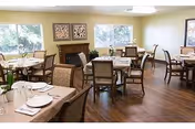 A dining room with several wooden tables and chairs arranged neatly. Each table is set with plates, napkins, and silverware. The room has large windows letting in natural light, a wooden floor, and a fireplace with decorative artwork above it.