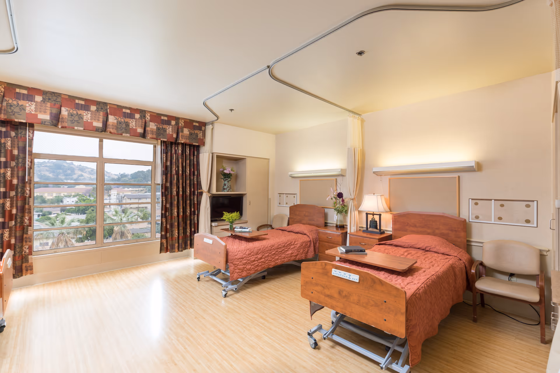 Shared patient bedroom with two adjustable beds, wooden bedside tables and lamps, chairs, and a large window with curtains showing a hillside view.