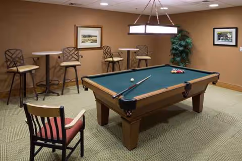A billiards table with stools, high tables, framed pictures, and a plant in beige-walled recreation room.