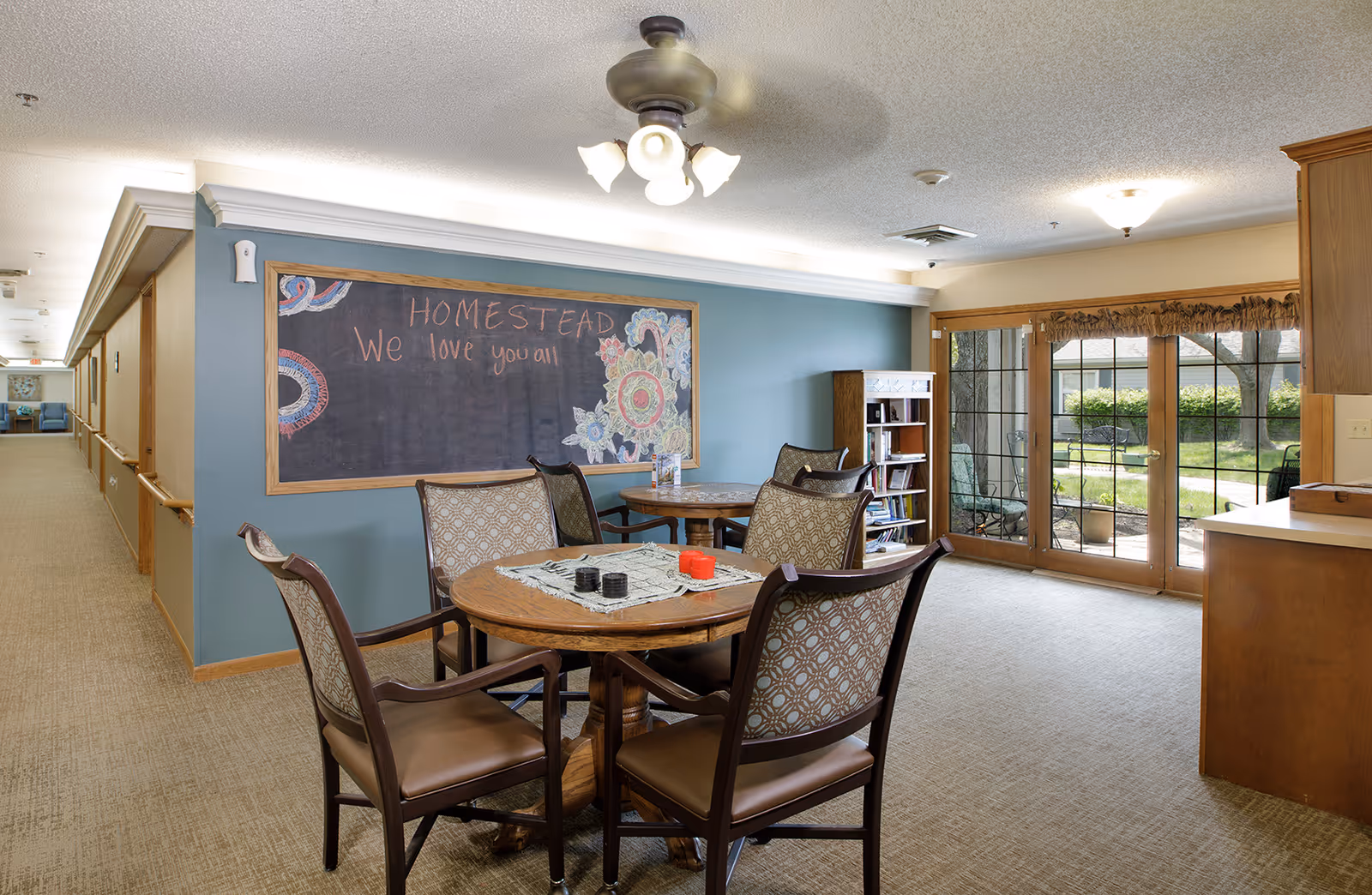 A cozy common area in Homestead Assisted Living of Olathe South featuring a round wooden table with four cushioned chairs. Behind the table is a large chalkboard on a blue wall with colorful chalk drawings and the message 'HOMESTEAD We love you all'. To the right, large glass doors open to a garden patio with outdoor seating. The room is well-lit with ceiling lights and has a carpeted floor.