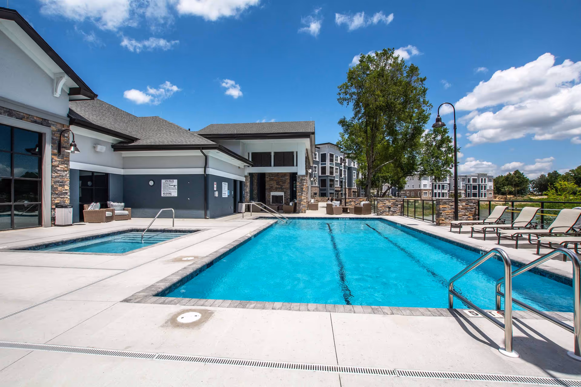 Outdoor swimming pool and hot tub area at Waterman Village Information Center with lounge chairs, seating area, and buildings in the background under a blue sky with some clouds.