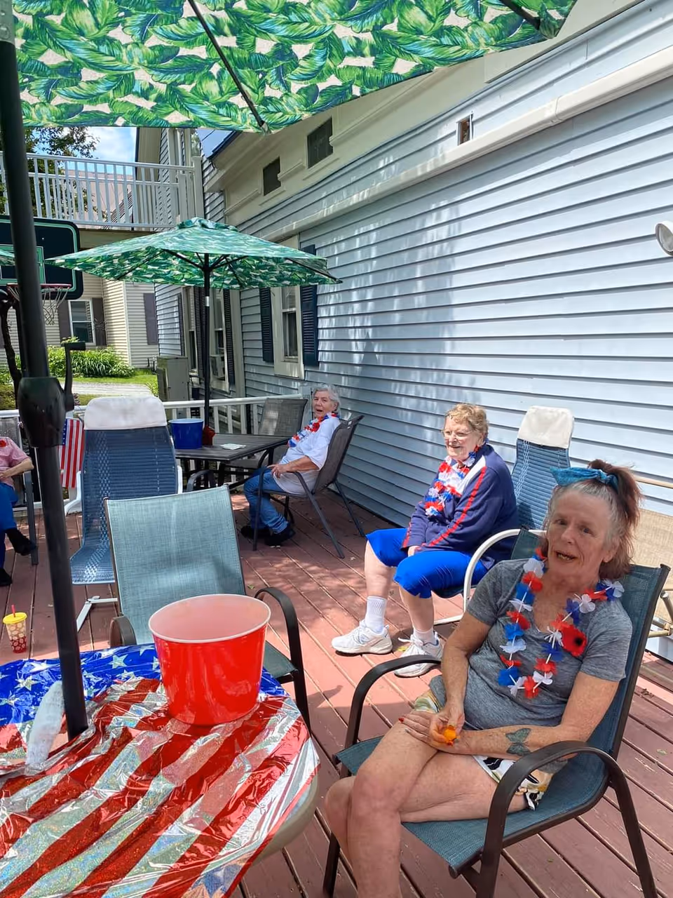 Three elderly women sitting on a wooden deck outside a light blue house. They are wearing red, white, and blue leis, suggesting a patriotic celebration. There are green umbrellas with leaf patterns providing shade, and a table covered with an American flag-themed tablecloth with a red bucket on it. The setting appears relaxed and social.