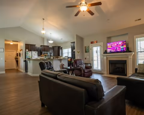 Spacious open-concept living area with dark leather sofas and a recliner facing a wall-mounted TV above a fireplace. The room has wood flooring, a ceiling fan with light, and large windows allowing natural light. The kitchen area with dark cabinets and a breakfast bar is visible in the background.