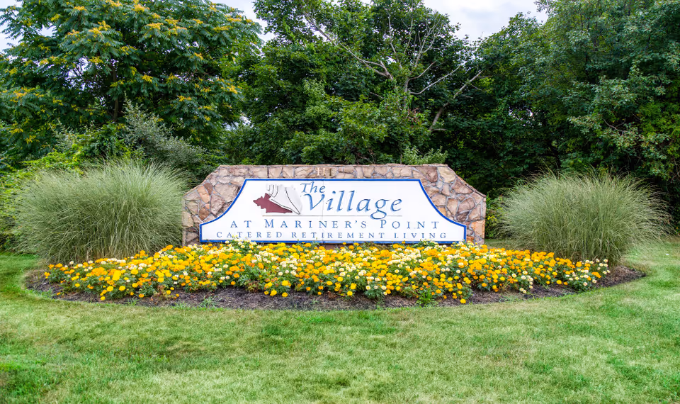 Stone entrance sign reading "The Village at Mariner's Point" surrounded by yellow flowers and landscaping.