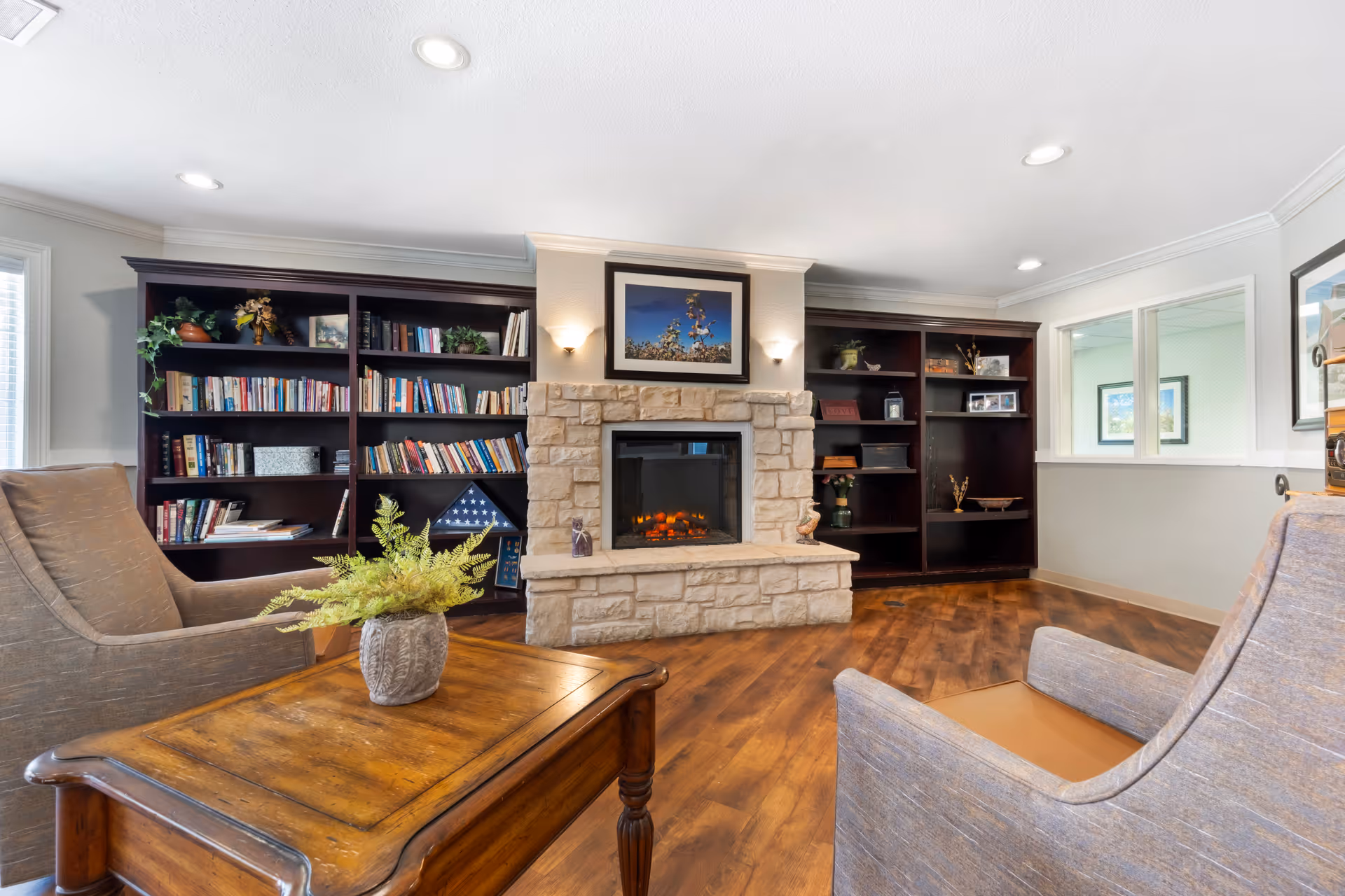 A cozy living room area with two upholstered armchairs facing a wooden coffee table with a potted plant. Behind the table is a stone fireplace with a framed picture above it, flanked by dark wooden bookshelves filled with books and decorative items. The room has wooden flooring and recessed ceiling lights.