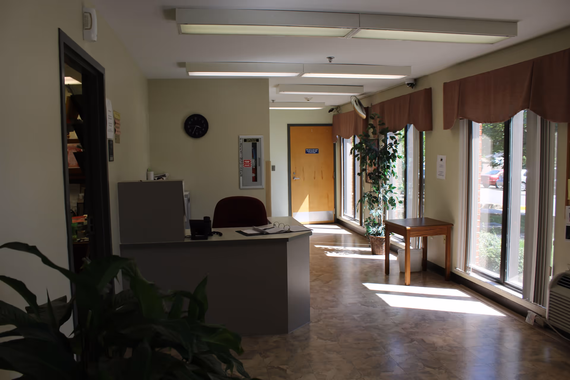 Interior view of a reception area with a desk and chair, a clock on the wall, a fire extinguisher cabinet, large windows with brown valances letting in sunlight, a potted plant, and a wooden table near the windows.