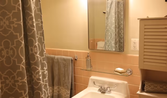 Bathroom sink with mirror above, pink tiled backsplash, a towel on a bar, patterned shower curtain, and a small wall cabinet.