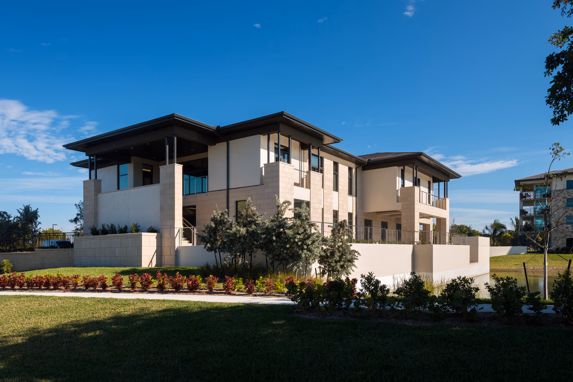 Modern two-story building with beige and light brown exterior walls, large windows, and a flat roof under a clear blue sky. The building is surrounded by landscaped greenery including bushes, trees, and a well-maintained lawn.