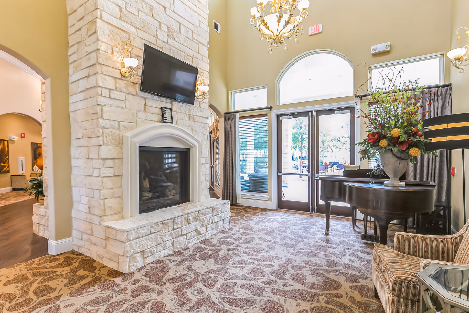 A bright and elegant living room area in Woodhaven Village featuring a large stone fireplace with a mounted flat-screen TV above it. The room has high ceilings with chandeliers, patterned carpet, a grand piano adorned with a large floral arrangement, and glass double doors leading outside. There is also a striped armchair and a side table in the foreground.