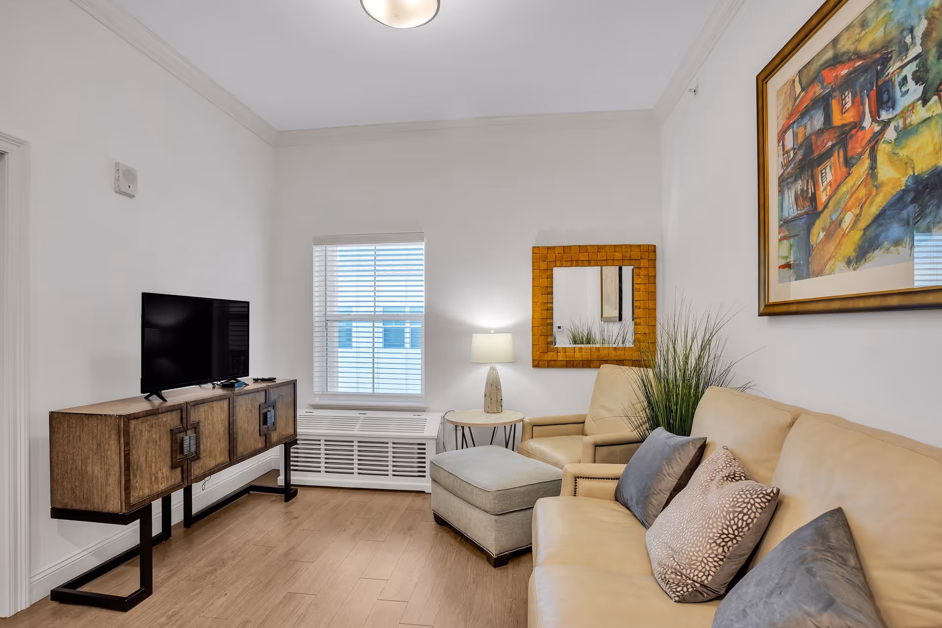 A cozy living room with a beige leather sofa adorned with decorative pillows, a matching armchair with an ottoman, a wooden TV stand with a flat-screen television, a small side table with a lamp, a window with blinds, a square mirror with a wooden frame, and a colorful framed painting on the wall.