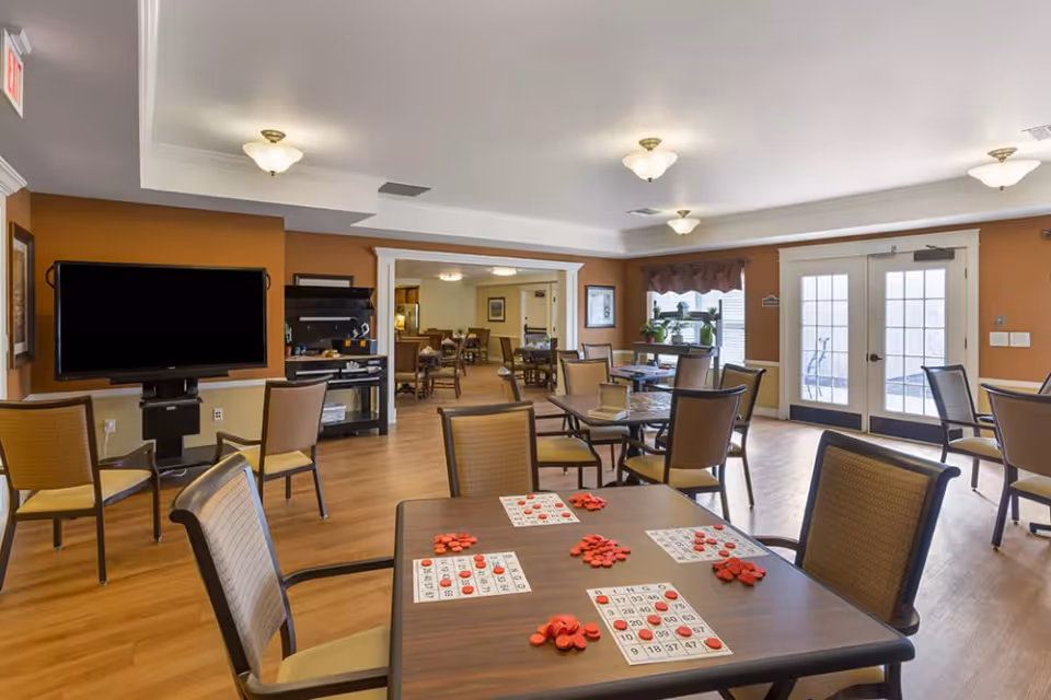 A spacious common area in an assisted living facility with multiple tables and chairs arranged for group activities. One table in the foreground has bingo cards and red markers on it. The room has warm brown walls, wood flooring, ceiling lights, a large flat-screen TV mounted on the wall, and double glass doors leading outside. There are additional seating areas and tables visible through a wide doorway in the background.