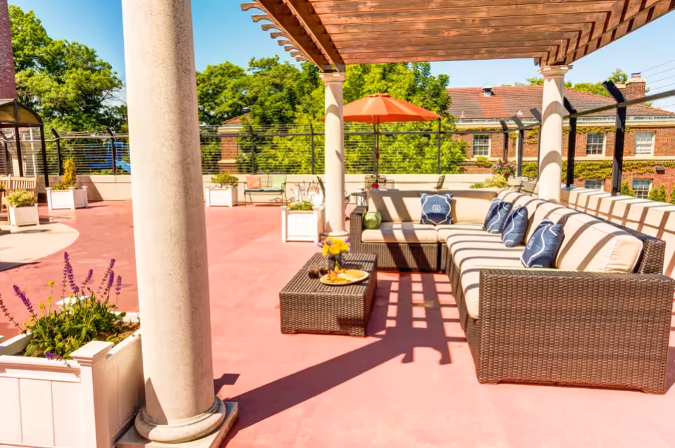 Outdoor patio area with a large wicker sectional sofa with beige cushions and blue pillows under a wooden pergola. There is a small wicker coffee table with a plate of food and a vase of flowers. The patio has pink flooring, white columns, potted plants, and an orange umbrella. In the background, there are trees and a brick building with windows.