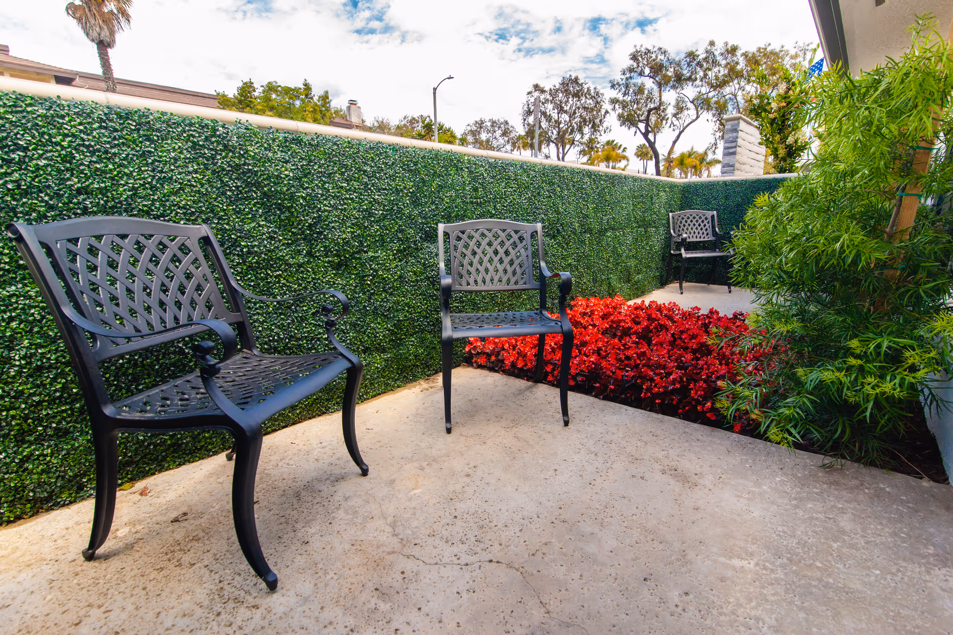 Outdoor patio area with three black metal chairs arranged around a small garden bed filled with red flowers. The patio is enclosed by a green hedge wall and surrounded by various plants and trees under a partly cloudy sky.