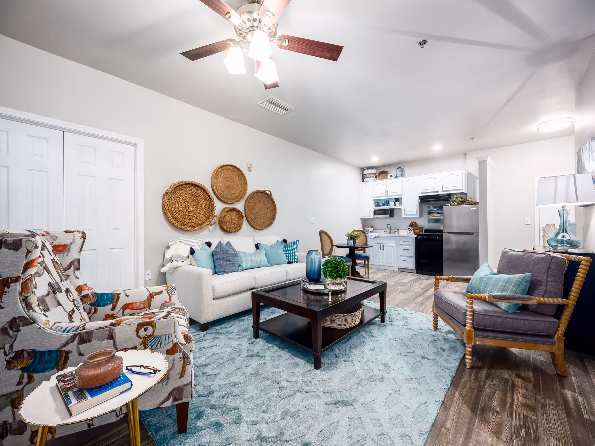 A bright and cozy living room area with a white sofa adorned with blue and teal pillows, a patterned armchair, and a wooden armchair with a teal cushion. A dark wooden coffee table sits on a light blue patterned rug. The wall behind the sofa features decorative woven baskets. In the background, there is a small kitchen with white cabinets, a black stove, and a stainless steel refrigerator. The room has wood flooring and a ceiling fan with lights.