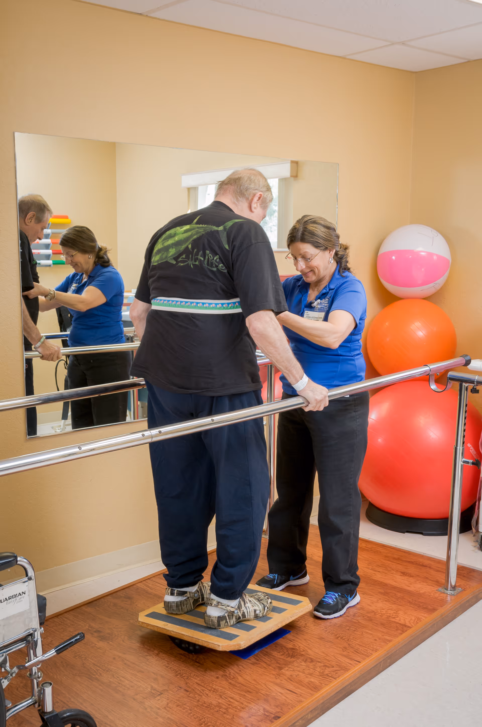 A senior man is standing on a balance board while holding onto parallel bars for support. A female therapist in a blue shirt is assisting him with the exercise in a therapy room with wooden floors, a large mirror, and colorful exercise balls in the background.