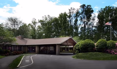 Front exterior view of Luther Woods Nursing & Rehabilitation Center showing a single-story building with a covered entrance, surrounded by trees and bushes, with an American flag on a flagpole to the right.