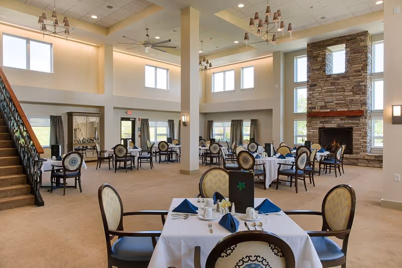 Spacious dining room with multiple round tables covered with white tablecloths, set with blue napkins, silverware, and cups. The room features large windows, a high ceiling with chandeliers, a stone fireplace, and a staircase with a decorative railing.