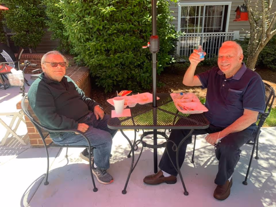 Two elderly men sitting at a black metal outdoor table with an umbrella pole in the center, enjoying a meal with disposable trays and cups. They are seated on metal chairs on a patio area with greenery and a building with windows in the background. One man is raising a small cup and smiling.
