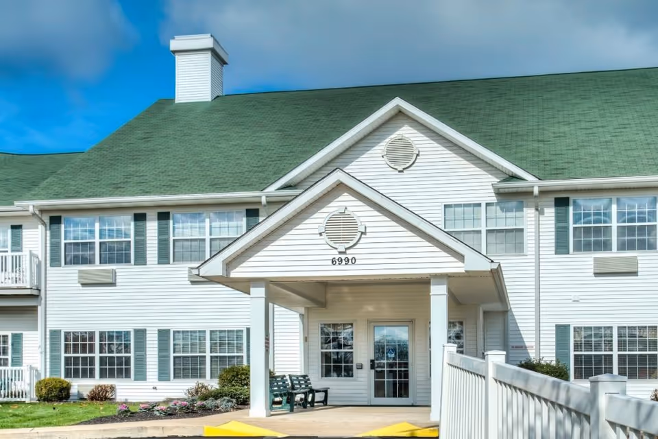 Front exterior view of a two-story senior living facility building with white siding and a green roof. The entrance has a covered porch with the number 6990 above it, a bench nearby, and a wheelchair accessible ramp leading to the door. The sky is clear with some clouds.