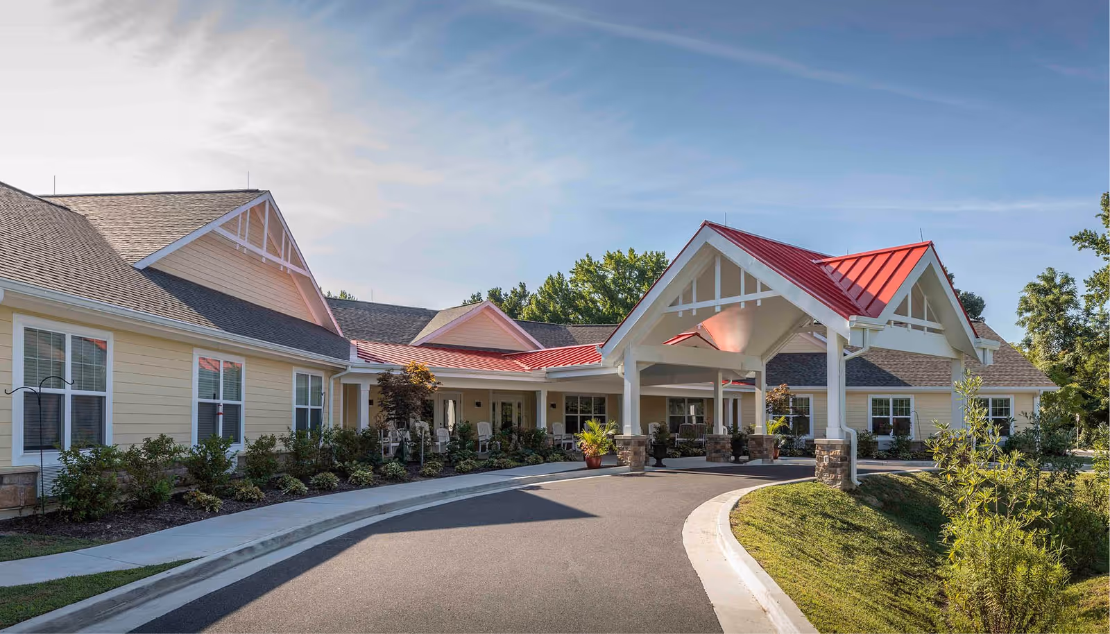 Exterior view of a single-story senior living facility building with a red metal roof over the entrance, beige siding, multiple windows, and a curved driveway leading to the covered entrance. There are shrubs and small trees planted along the building and a clear blue sky above.