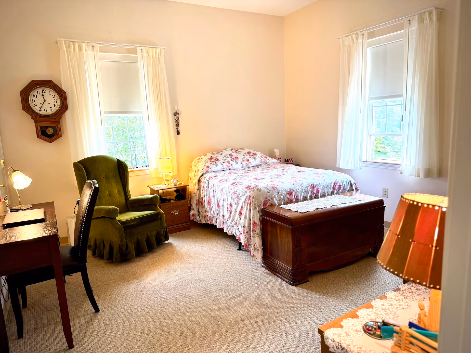 Well-lit furnished bedroom with a floral bedspread, a green armchair, desk, and wooden chest at the foot of the bed.