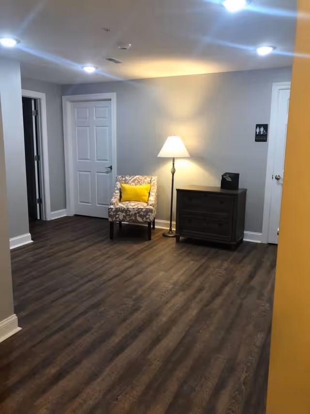A hallway area with wood flooring, a patterned armchair with a yellow cushion, a floor lamp, and a dark wooden cabinet. There are two white doors, one on the left and one on the right, with a restroom sign on the wall next to the right door.