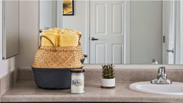 Bathroom countertop with a sink, a woven basket filled with yellow towels, a small potted plant, and a candle. A large mirror reflects a white door and part of the bathroom.