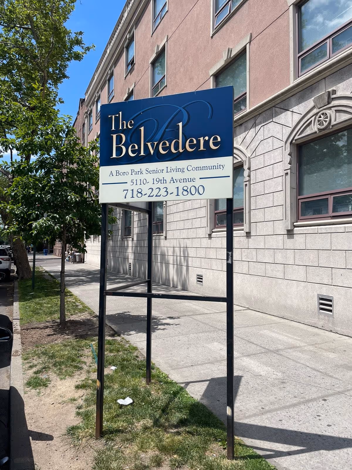 A sidewalk view of a sign for The Belvedere, a Boro Park Senior Living Community, located at 5110-19th Avenue with the phone number 718-223-1800. The sign is mounted on black metal poles and is positioned next to a building with stone and brick exterior walls and several windows. There is a tree and some grass along the sidewalk under a clear blue sky.