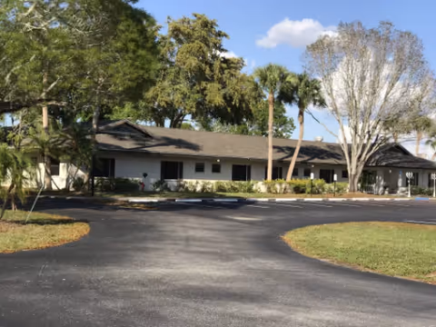 Single-story light-colored building with a low roof set behind a curved asphalt driveway, surrounded by palm trees and landscaping under a blue sky.