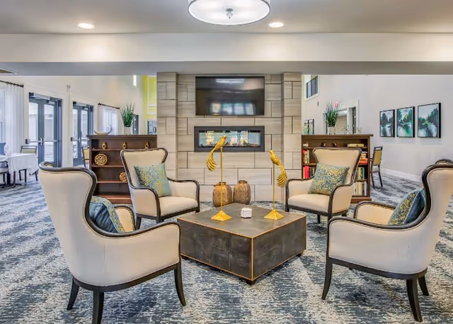 Well-lit communal lounge with four upholstered armchairs arranged around a low coffee table facing a wall-mounted TV and fireplace.