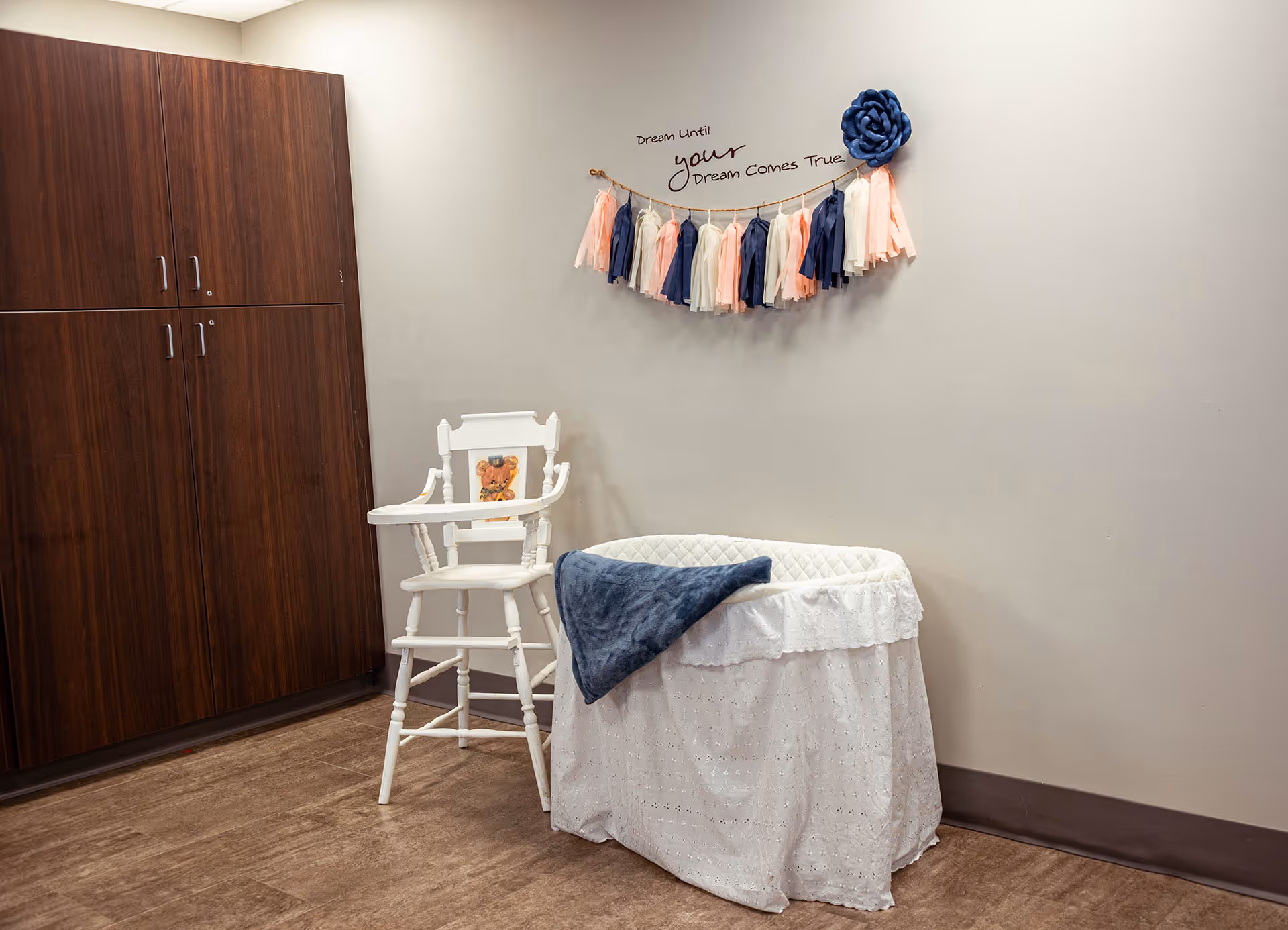 Corner of an interior room with a white lace-covered bassinet, a white high chair, a decorative wall garland, and dark wood storage cabinets.