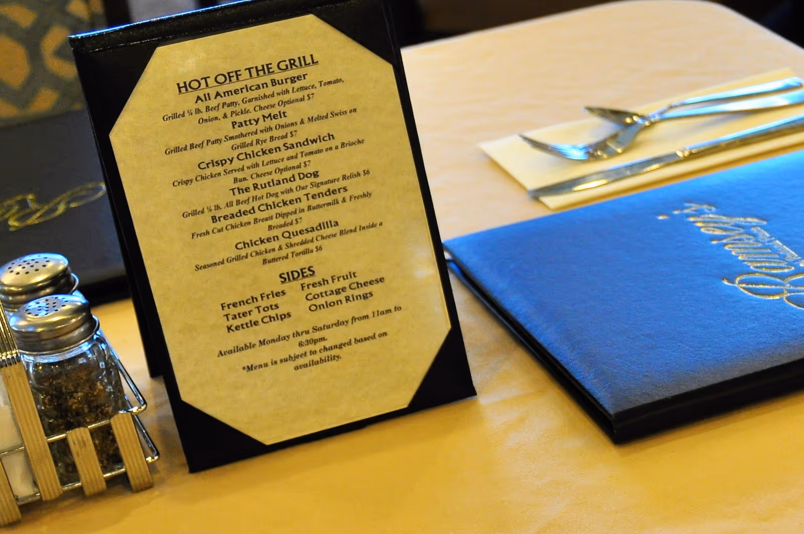 A close-up view of a table in a dining area featuring a menu titled 'Hot Off The Grill' with various food options listed, including burgers, sandwiches, and sides. The table also has salt and pepper shakers, a blue menu folder, and silverware wrapped in a napkin.