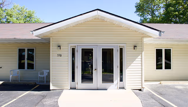 Front entrance of a single-story senior living facility with double doors and seating outside