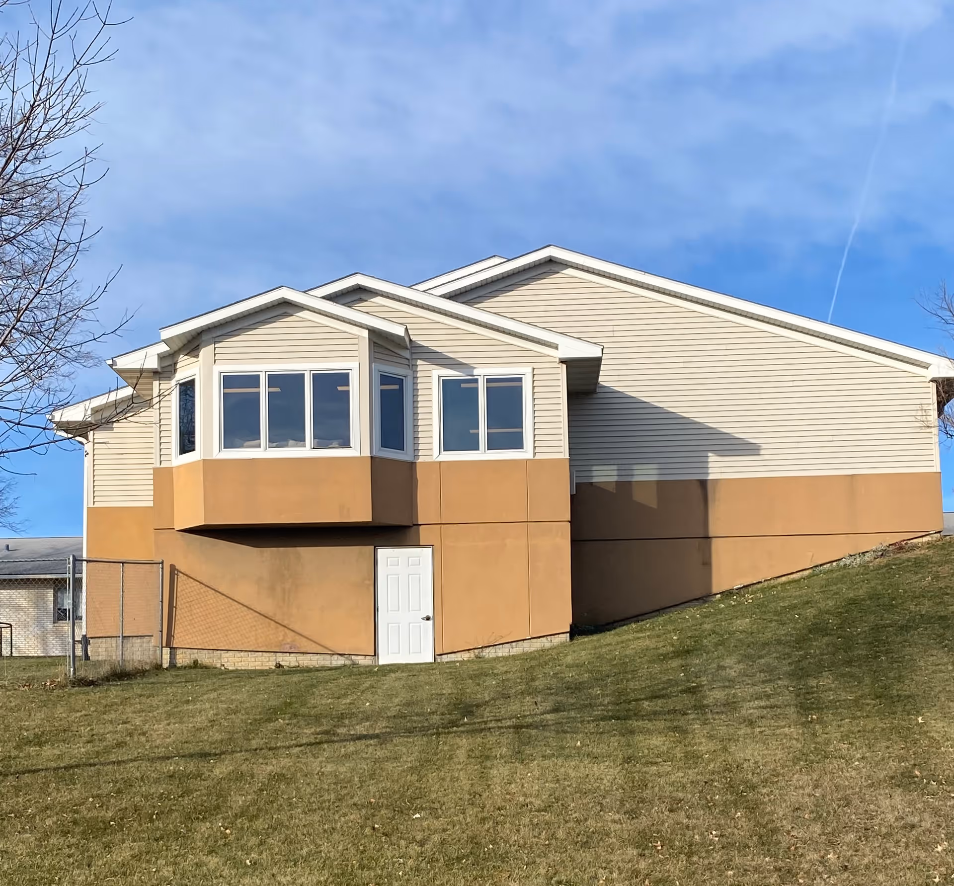 Exterior view of a beige and brown building with a white door and several windows, situated on a grassy slope under a blue sky.