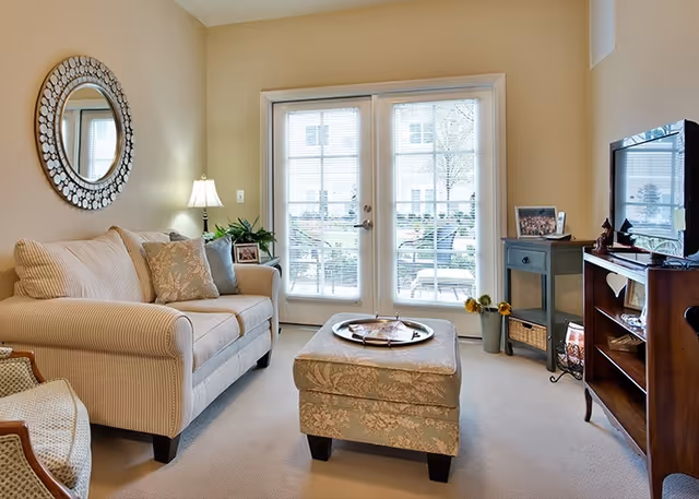 A cozy living room with a beige striped sofa adorned with decorative pillows, a round mirror on the wall, a floor lamp, and a cushioned ottoman with a tray on top. French doors with blinds lead to an outdoor area. There is a wooden TV stand with a flat-screen television and a small side table with framed photos and a vase with sunflowers.
