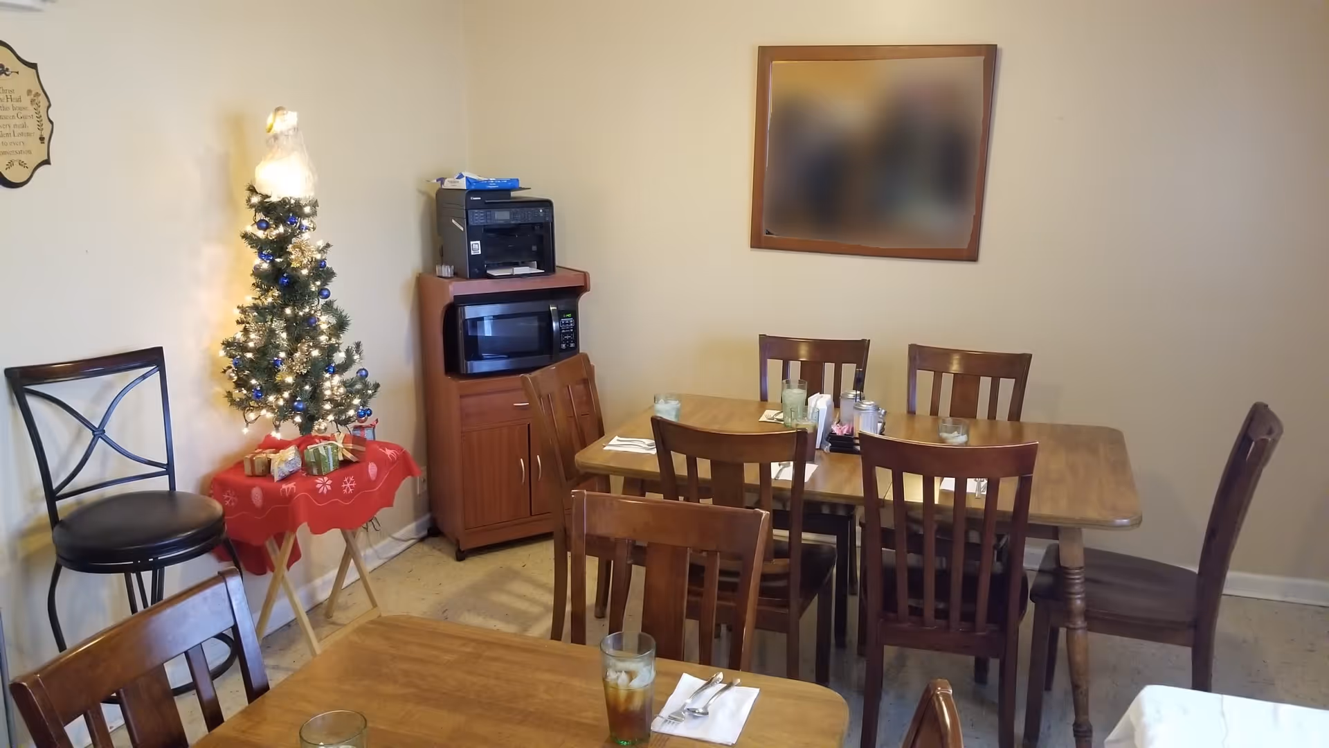 A small dining area with wooden tables and chairs. One table has glasses of iced tea and napkins with utensils. In the corner, there is a small decorated Christmas tree on a red tablecloth-covered table. A wooden cabinet holds a microwave and a printer. A framed picture is on the wall above the tables.