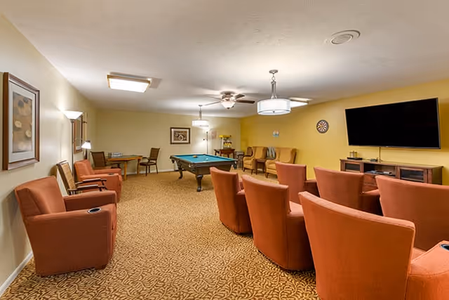 A spacious common room with several orange upholstered chairs arranged in rows facing a large flat-screen TV mounted on a yellow wall. The room also features a pool table in the center, a dartboard on the wall, and additional seating and tables along the sides. The carpet has a patterned design, and the ceiling has multiple light fixtures including ceiling fans.