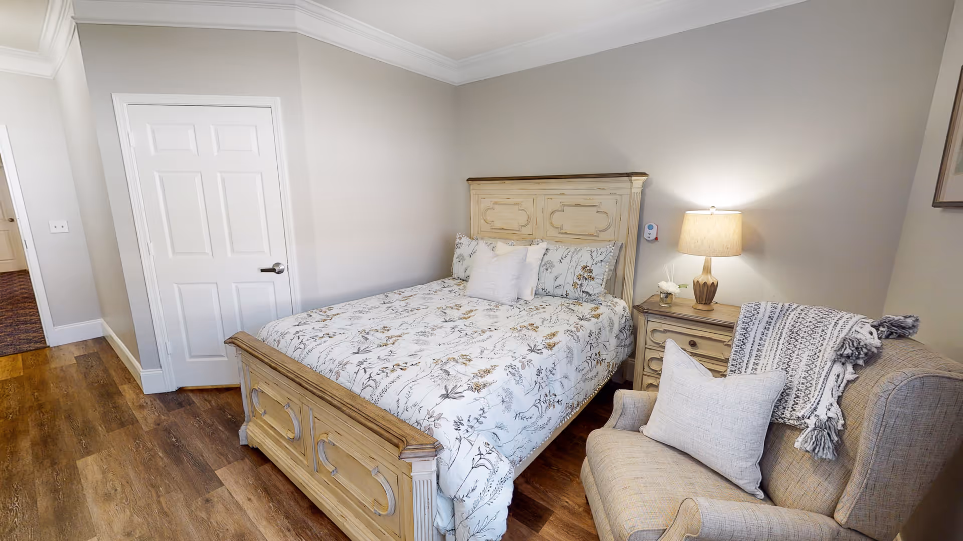 A cozy bedroom with a wooden bed frame and floral patterned bedding. Next to the bed is a wooden nightstand with a lamp and a small flower vase. A comfortable armchair with a pillow and a throw blanket is positioned near the nightstand. The room has wooden flooring and light-colored walls with a closed white door visible.