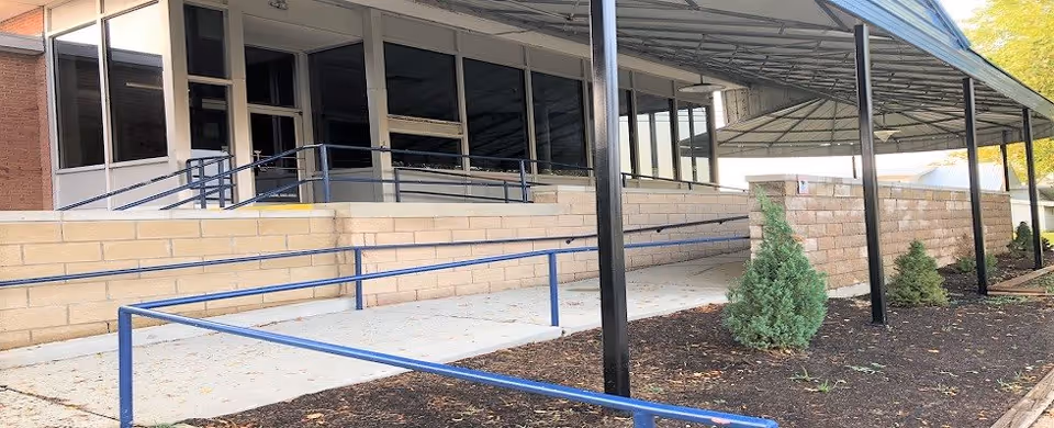 Exterior view of a building entrance with a covered walkway and blue handrails along a concrete ramp. There are small evergreen shrubs planted in mulch beds beside the ramp and a brick wall supporting the walkway.