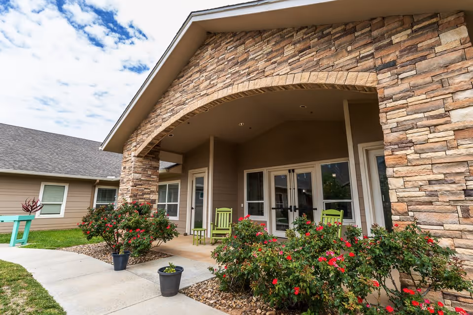 Exterior view of a senior living facility entrance with stone archway, beige siding, green rocking chairs on the porch, and flowering bushes along the walkway under a partly cloudy sky.