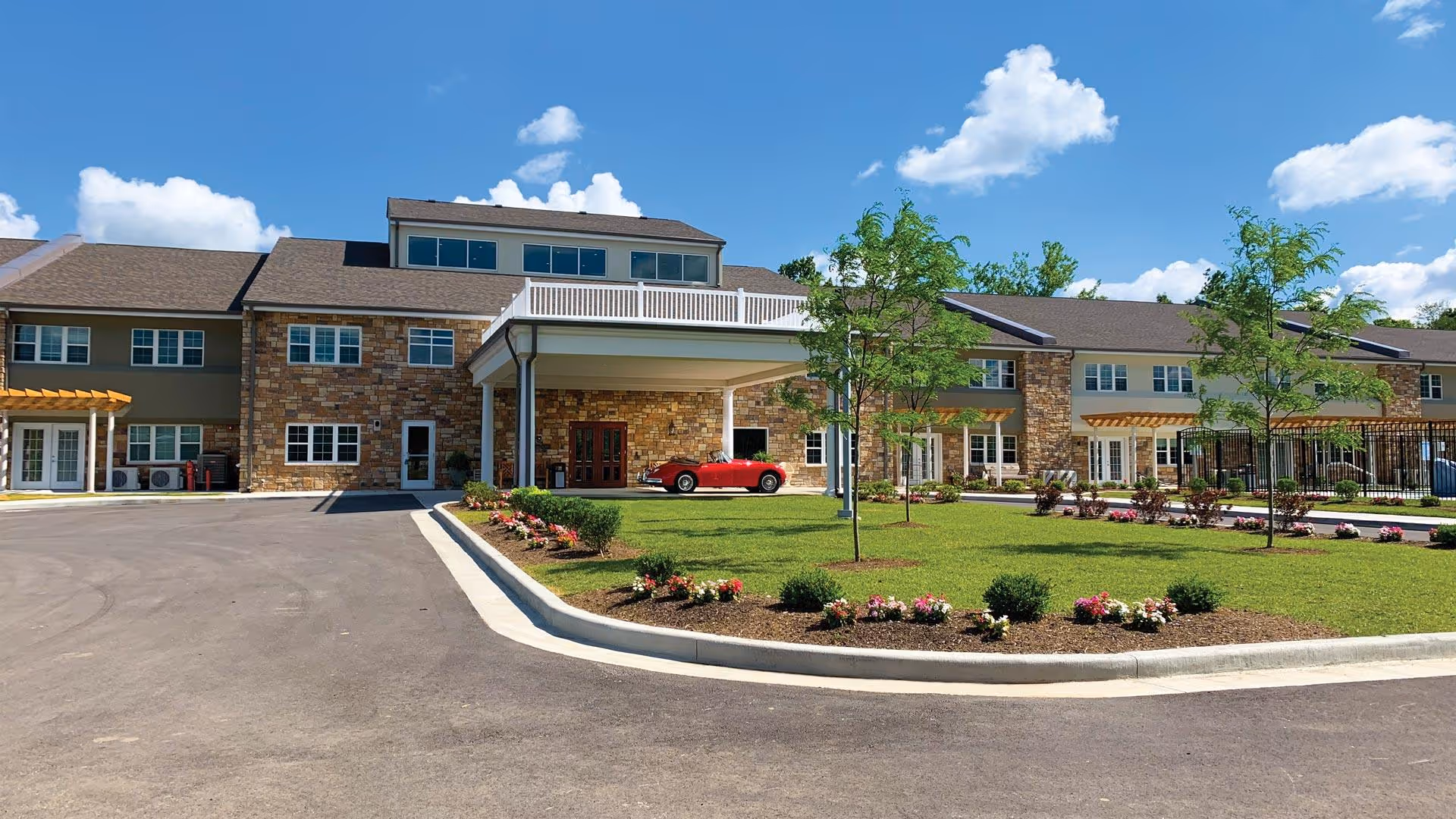 Exterior view of Sycamore Reserve Senior Living facility showing a two-story building with stone and beige siding, a covered entrance with a red car parked underneath, landscaped lawn with small trees and flower beds, and a clear blue sky with scattered clouds.