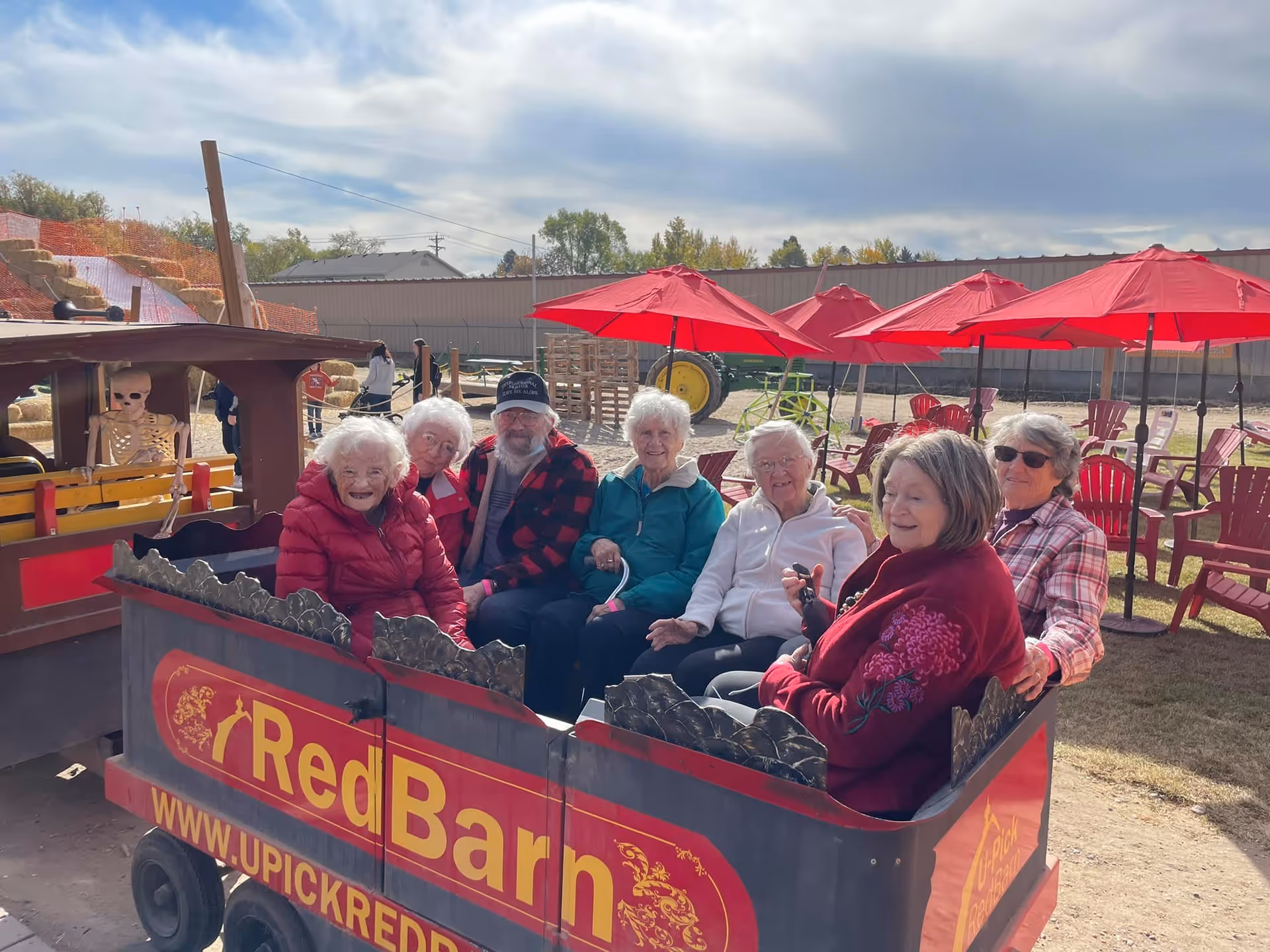 A group of seven elderly people sitting together in a small open wagon labeled 'Red Barn' with a website URL www.upickredbarn.com. They are outdoors on a sunny day with red umbrellas and chairs in the background. A skeleton decoration is visible on the left side of the wagon.
