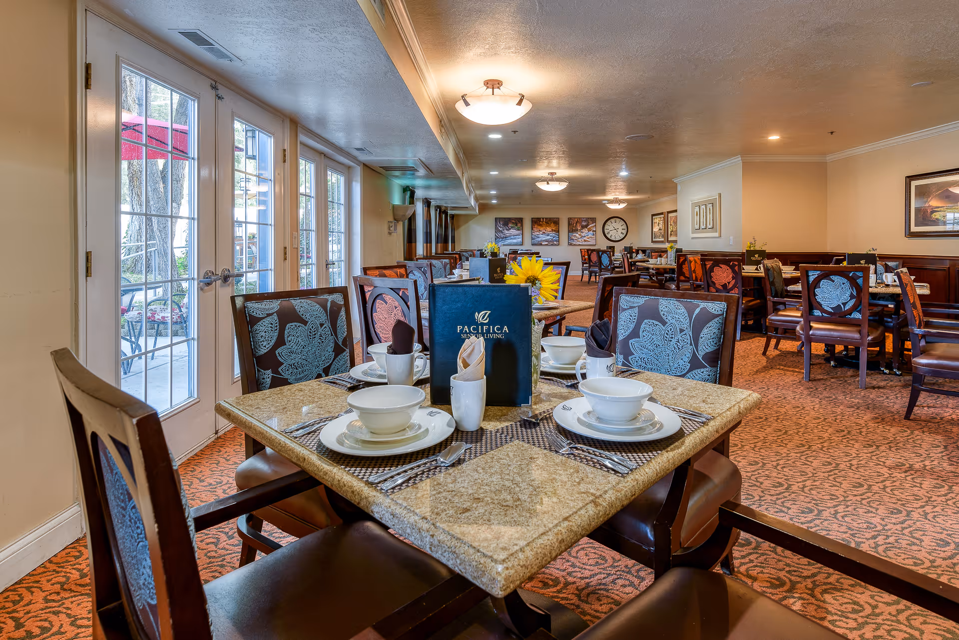 Interior view of a senior living facility dining room with multiple tables and chairs. A table in the foreground is set with white bowls, plates, cups, silverware, and napkins, along with a menu labeled 'Pacifica Senior Living' and a vase with a yellow sunflower. Large windows and glass doors on the left side let in natural light, and the room has warm lighting and carpeted floors with patterned upholstery on the chairs.