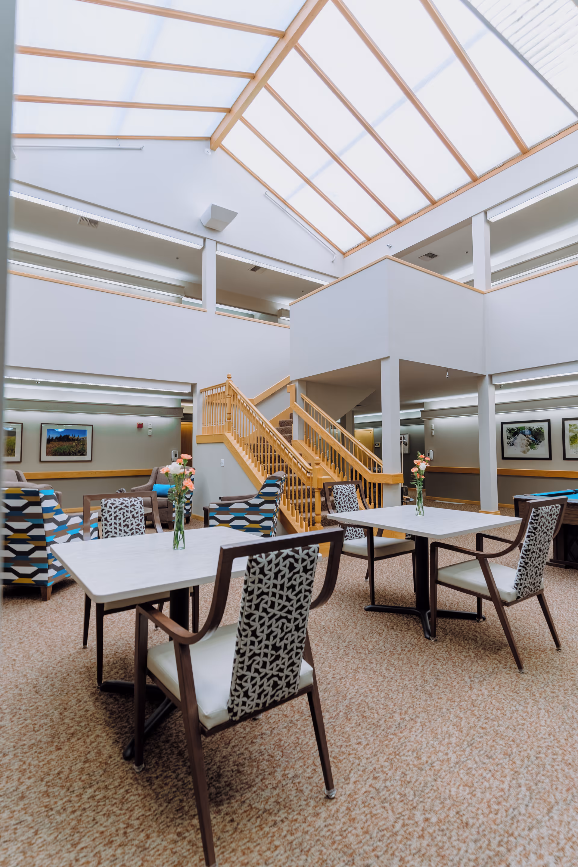 Bright and spacious common area in a senior living facility with patterned chairs around tables, small vases with flowers on the tables, a wooden staircase leading to an upper level, and a large skylight ceiling allowing natural light to fill the space.