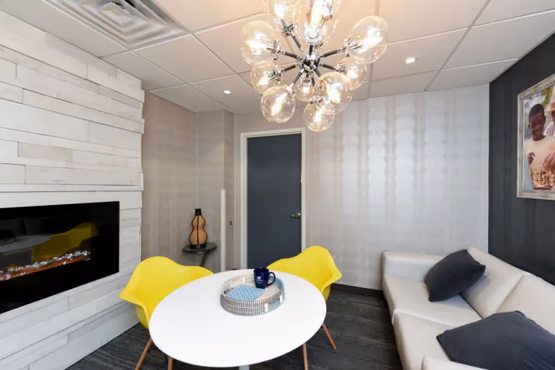 A modern interior room featuring a white round table with a decorative tray and a blue mug on top, surrounded by two bright yellow chairs. To the right is a white sofa with dark cushions. The wall behind the sofa has a framed picture of two children playing chess. On the left wall, there is a sleek black electric fireplace set into a white stone wall. A contemporary chandelier with multiple glass globes hangs from the ceiling.