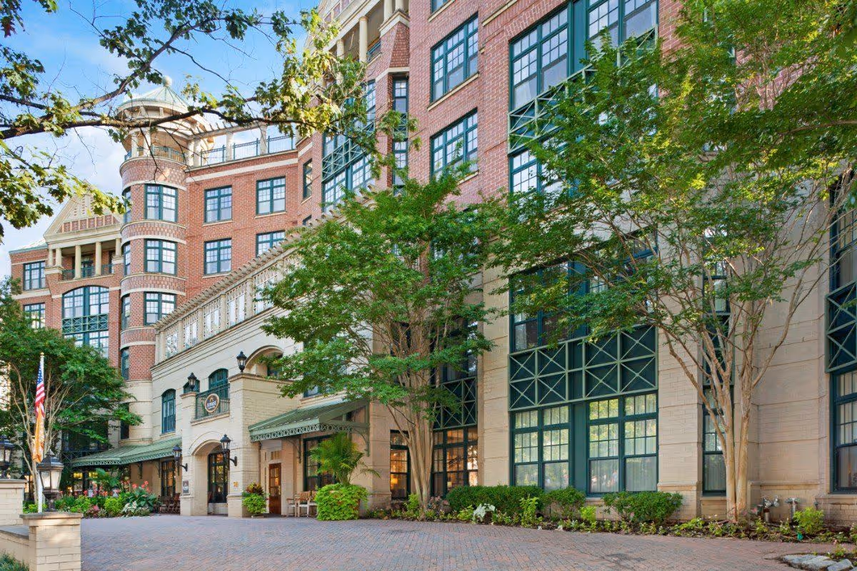 Exterior view of a multi-story red-brick building with large green-framed windows, trees, and a paved entrance.