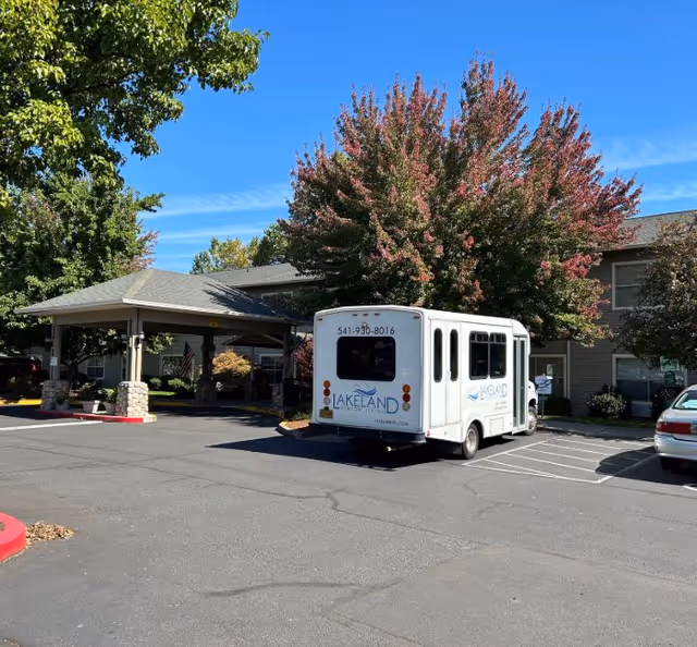 Exterior view of Lakeland Senior Living facility parking lot with a white shuttle bus parked near the entrance. The building is surrounded by trees with autumn foliage under a clear blue sky.