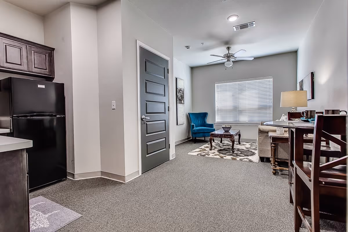 Interior view of a senior living facility unit showing a small living area with a blue armchair, a coffee table on a patterned rug, a sofa, a side table with a lamp, and a dining table with chairs. To the left, there is a kitchen area with dark wooden cabinets and a black refrigerator. A closed gray door is visible in the center of the image.